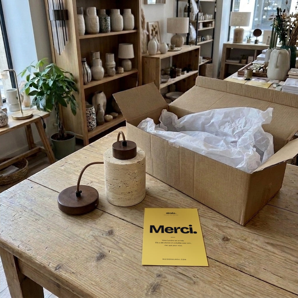 Suspension cylindre en travertin et rosace noyer, déballée sur la table de la boutique avec la carte Merci