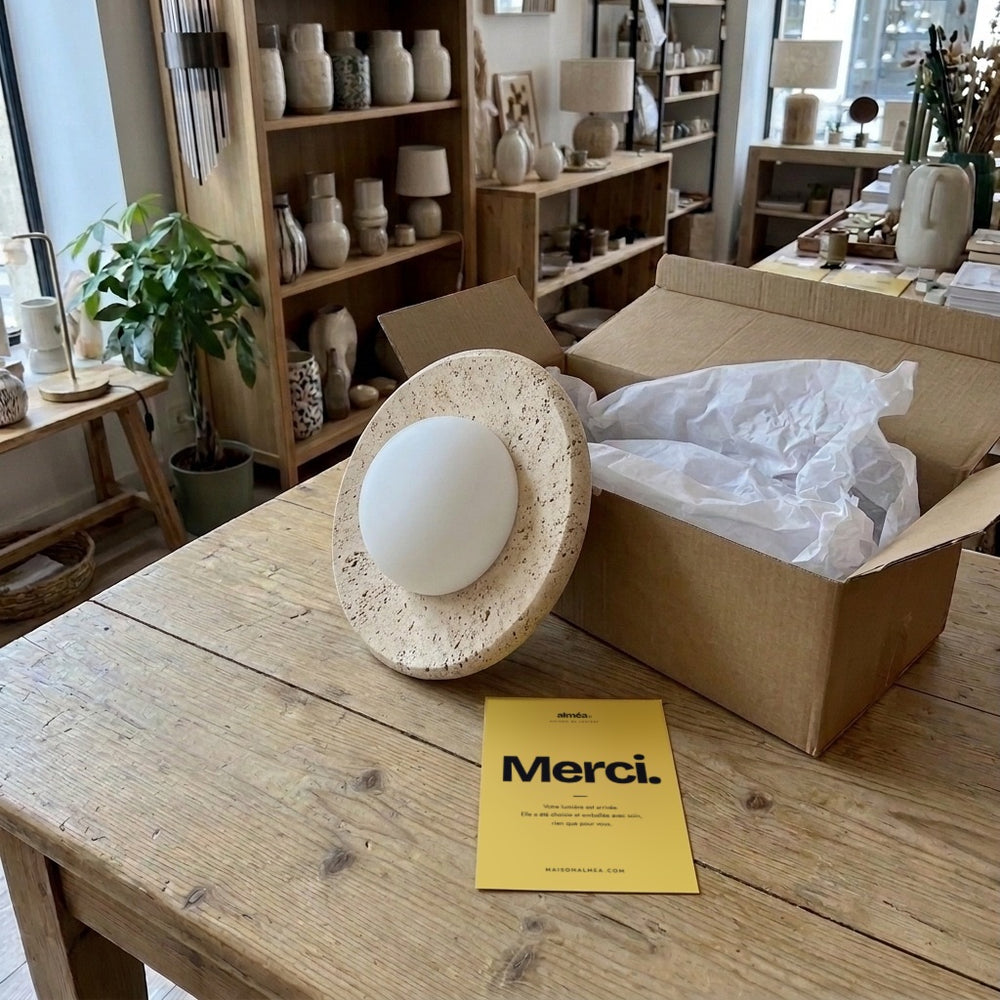 Plafonnier en travertin, globe opalin, déballé sur la table de la boutique avec la carte Merci