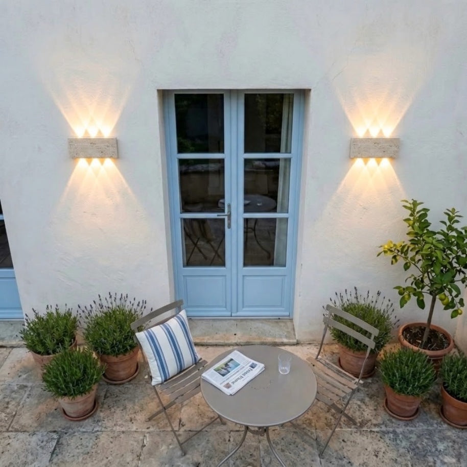 Deux appliques en travertin allumées de part et d'autre d'une porte-fenêtre bleue, terrasse provençale le soir