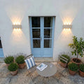 Deux appliques en travertin allumées de part et d'autre d'une porte-fenêtre bleue, terrasse provençale le soir